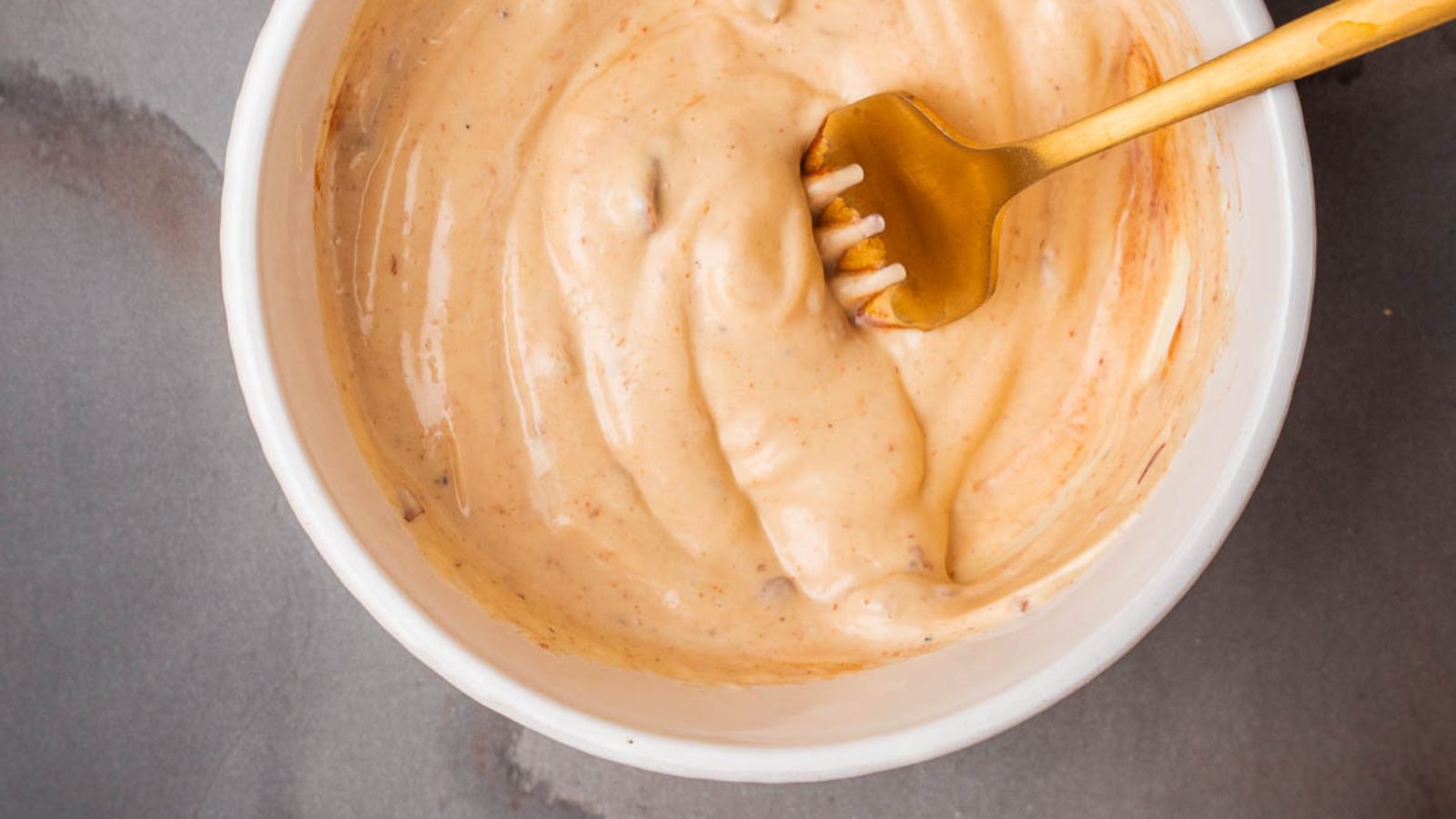 A gold fork rests in a white bowl filled with a creamy, light orange sauce on a gray surface.