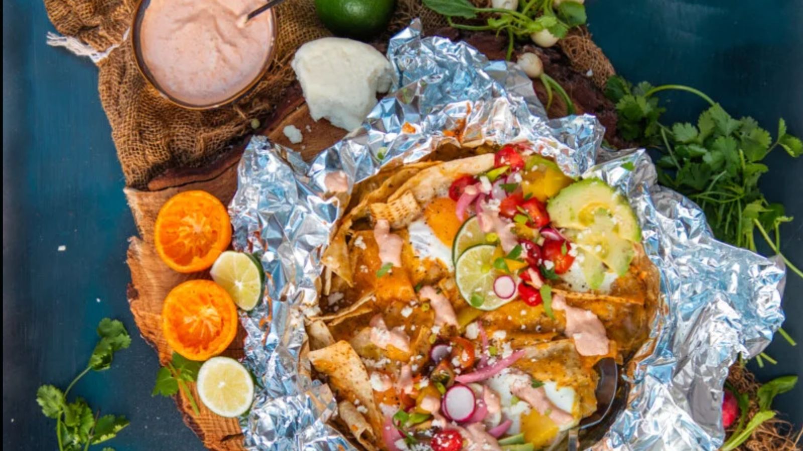 A foil tray of loaded nachos topped with avocado, radish, onions, pomegranate seeds, and sauce, surrounded by citrus halves, herbs, cheese, and a bowl of dip on a wooden board.