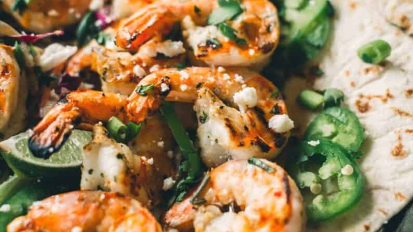 Grilled shrimp garnished with chopped herbs, sliced jalapeños, crumbled cheese, and lime wedges on a tortilla.
