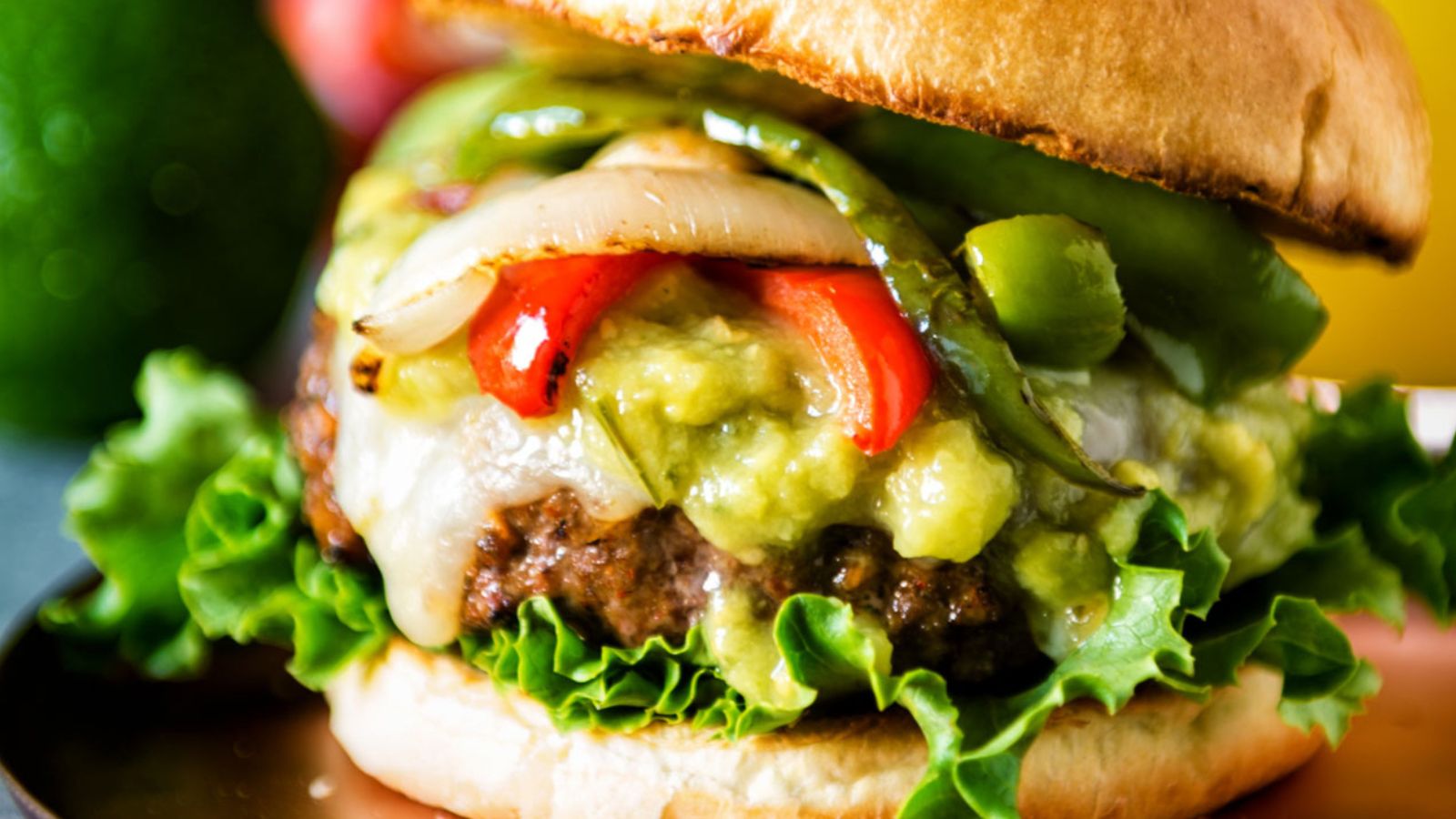 A close-up of a hamburger with lettuce, melted cheese, grilled onions and peppers, guacamole, and a toasted bun.