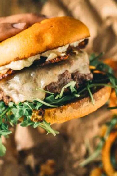 A hand holds a hamburger with melted cheese, bacon, lettuce, and arugula in a bun. Onion rings and greens are visible on the brown paper background.