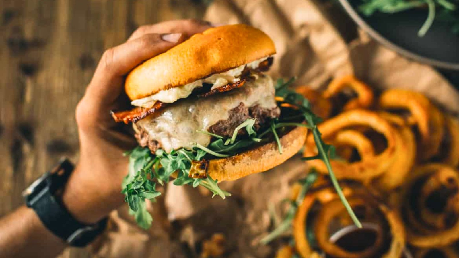 A hand holds a hamburger with melted cheese, bacon, lettuce, and arugula in a bun. Onion rings and greens are visible on the brown paper background.