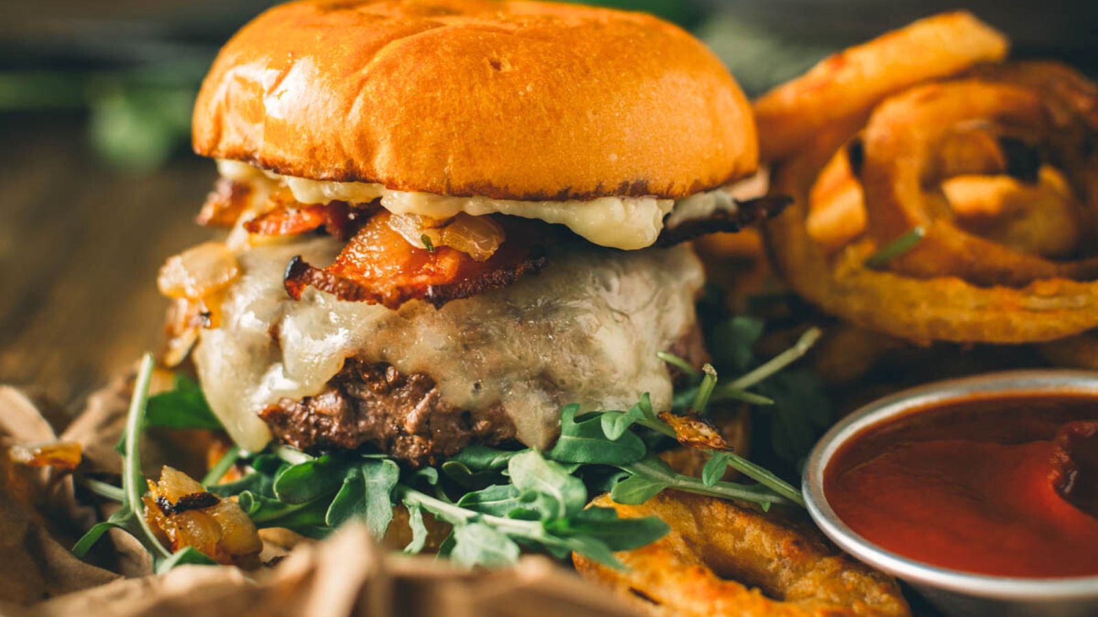 A cheeseburger with bacon, lettuce, and onions on a bun, served with onion rings and a side of ketchup.
