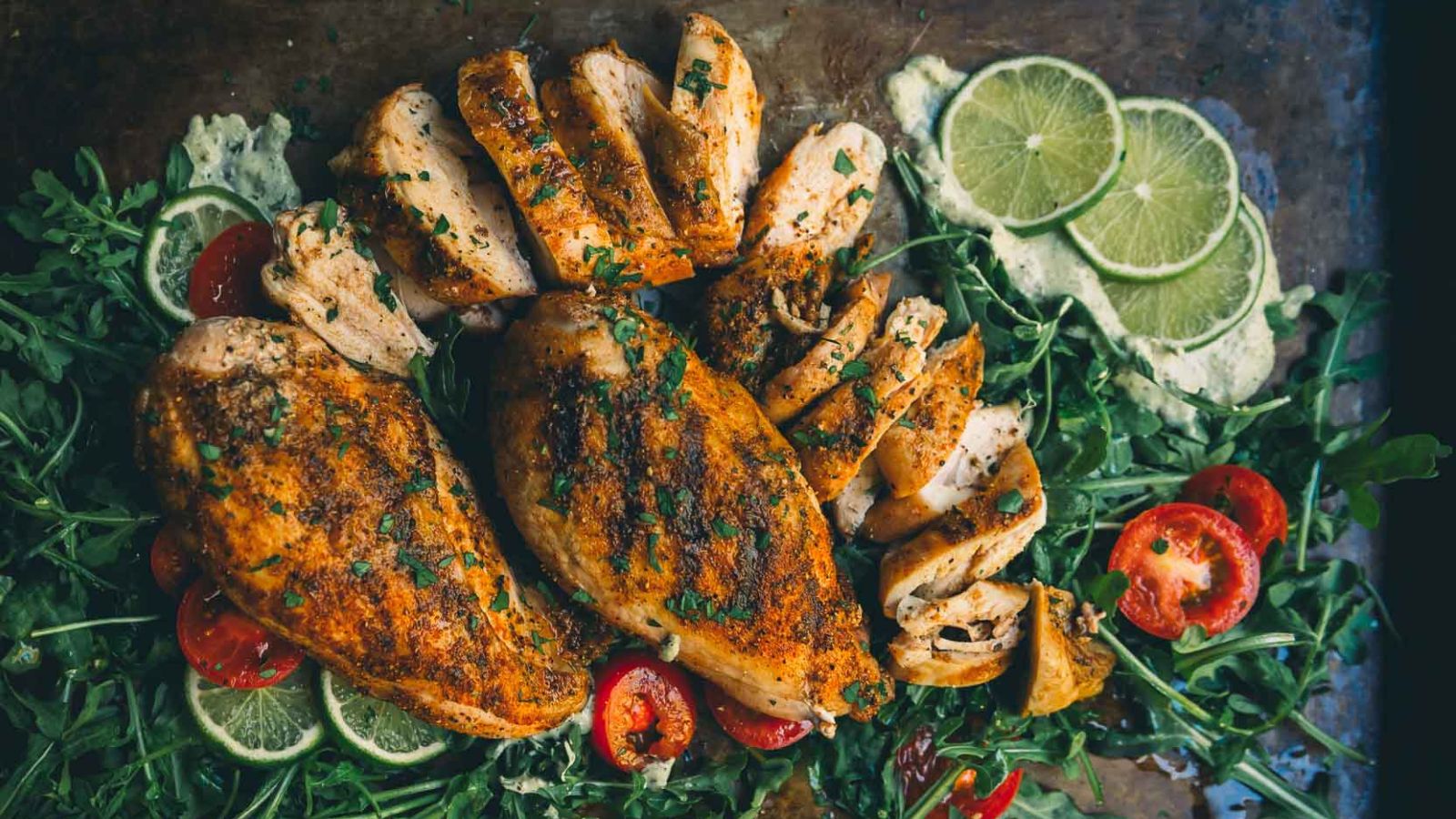Sliced, seasoned grilled chicken breasts served on a bed of greens with lime slices and cherry tomatoes.