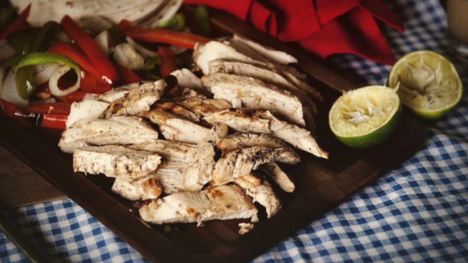Sliced grilled chicken breast on a wooden board, with mixed sautéed peppers and onions, tortillas, and halved limes on a blue checkered tablecloth.