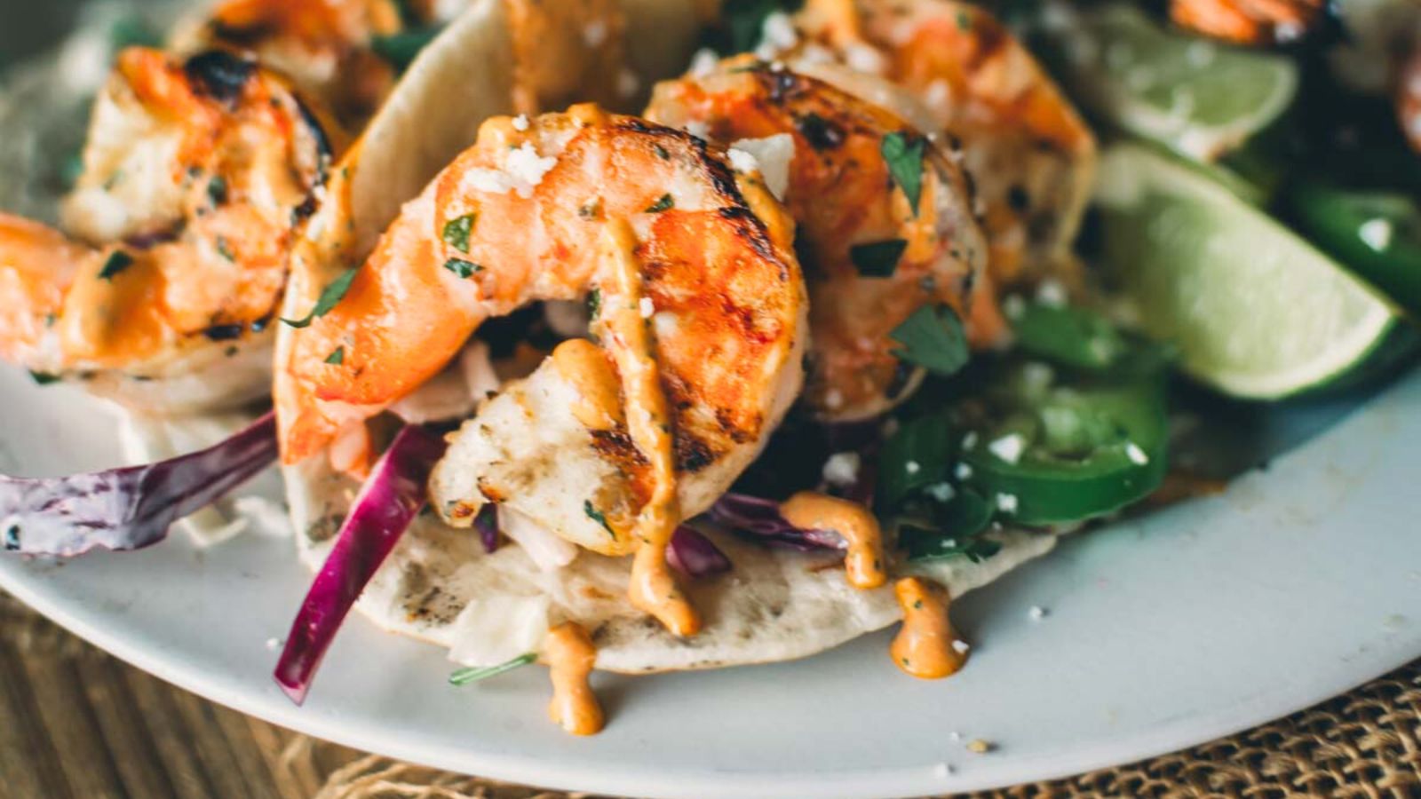 Grilled shrimp tacos with red cabbage, sliced jalapeños, cilantro, creamy sauce, and lime wedges on a white plate.
