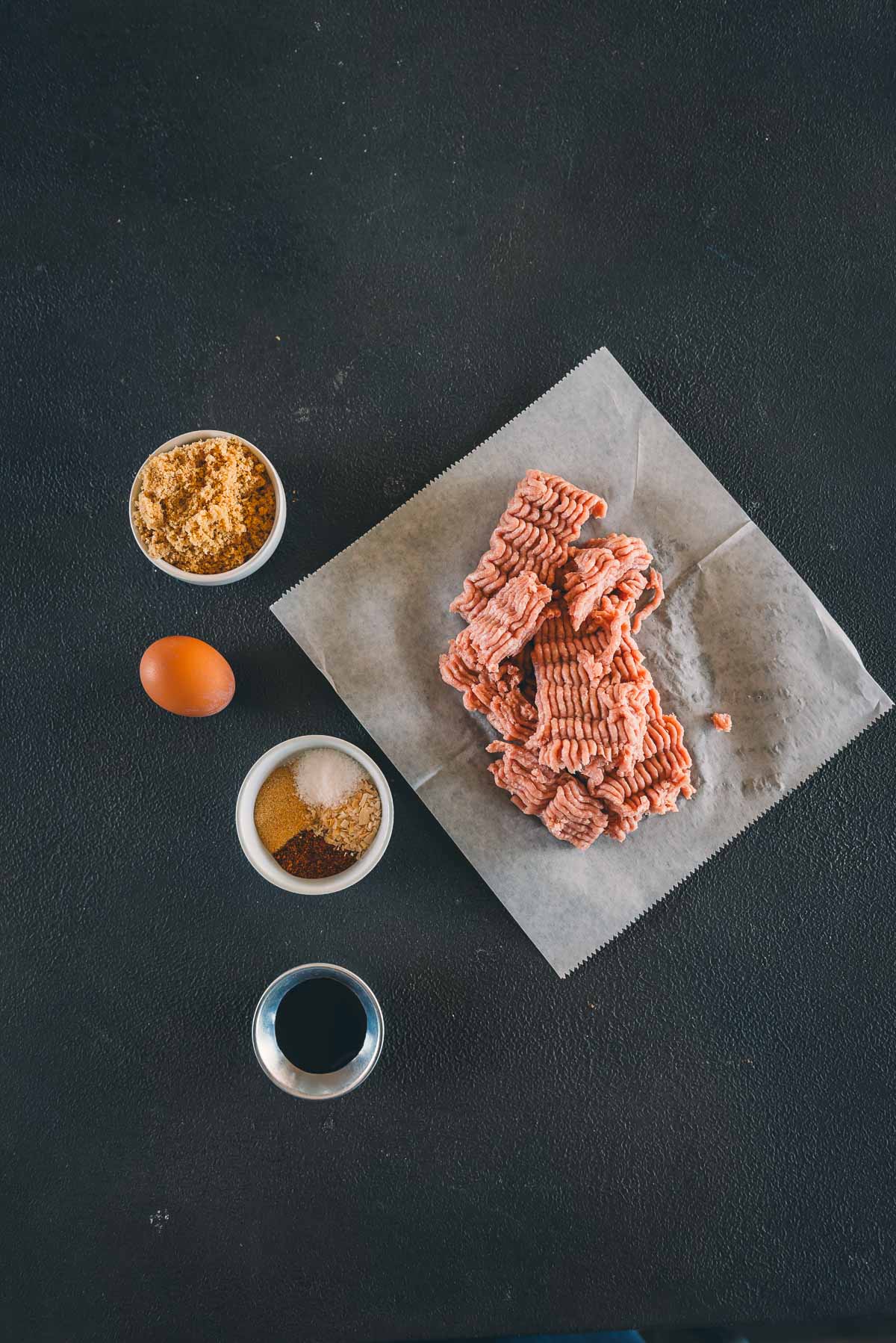 Ground turkey on parchment paper, surrounded by small bowls containing brown sugar, an egg, spices, and a dark liquid, all on a dark surface.