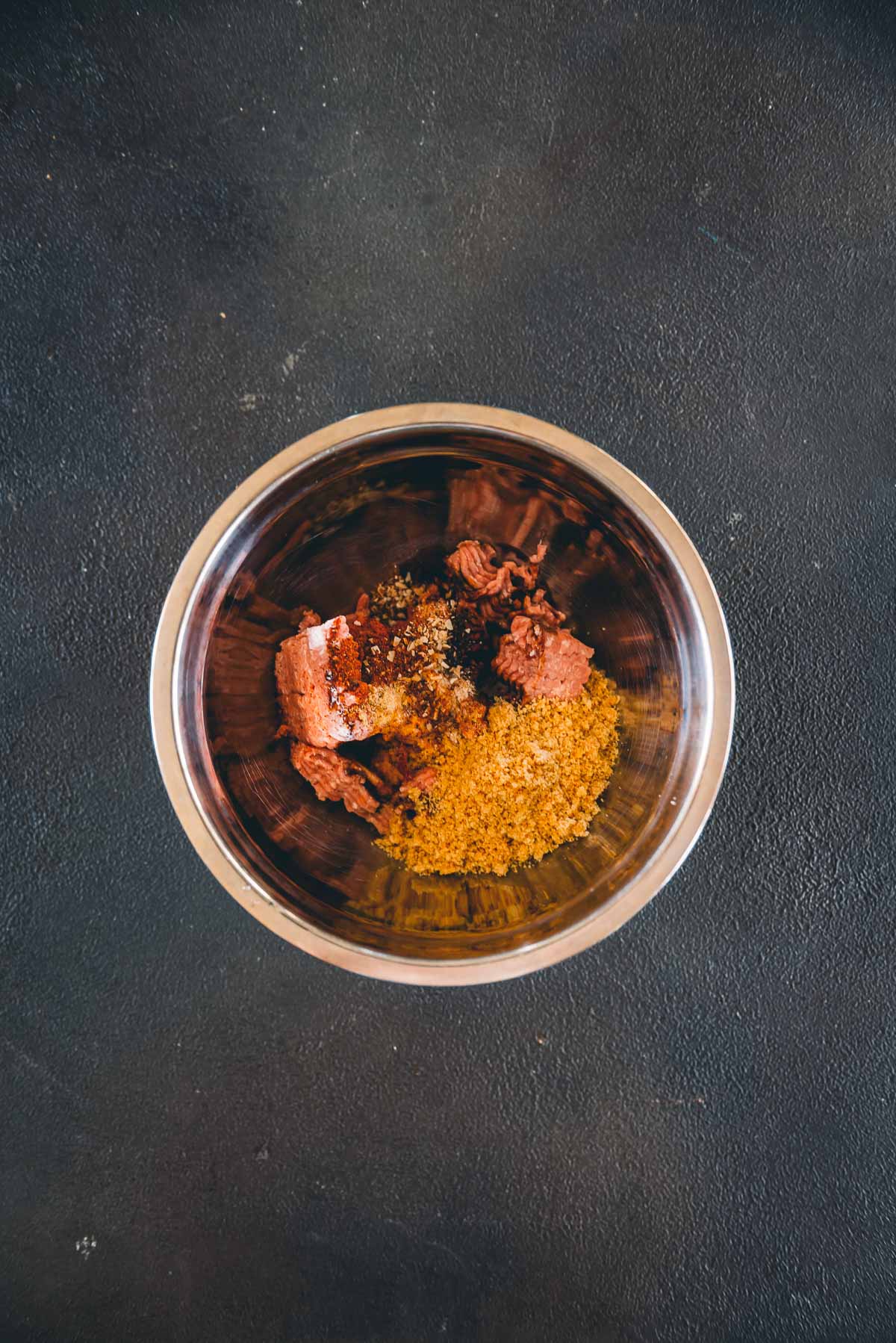 A metal bowl containing various ground spice, seasonings and ground turkey on a dark textured surface.