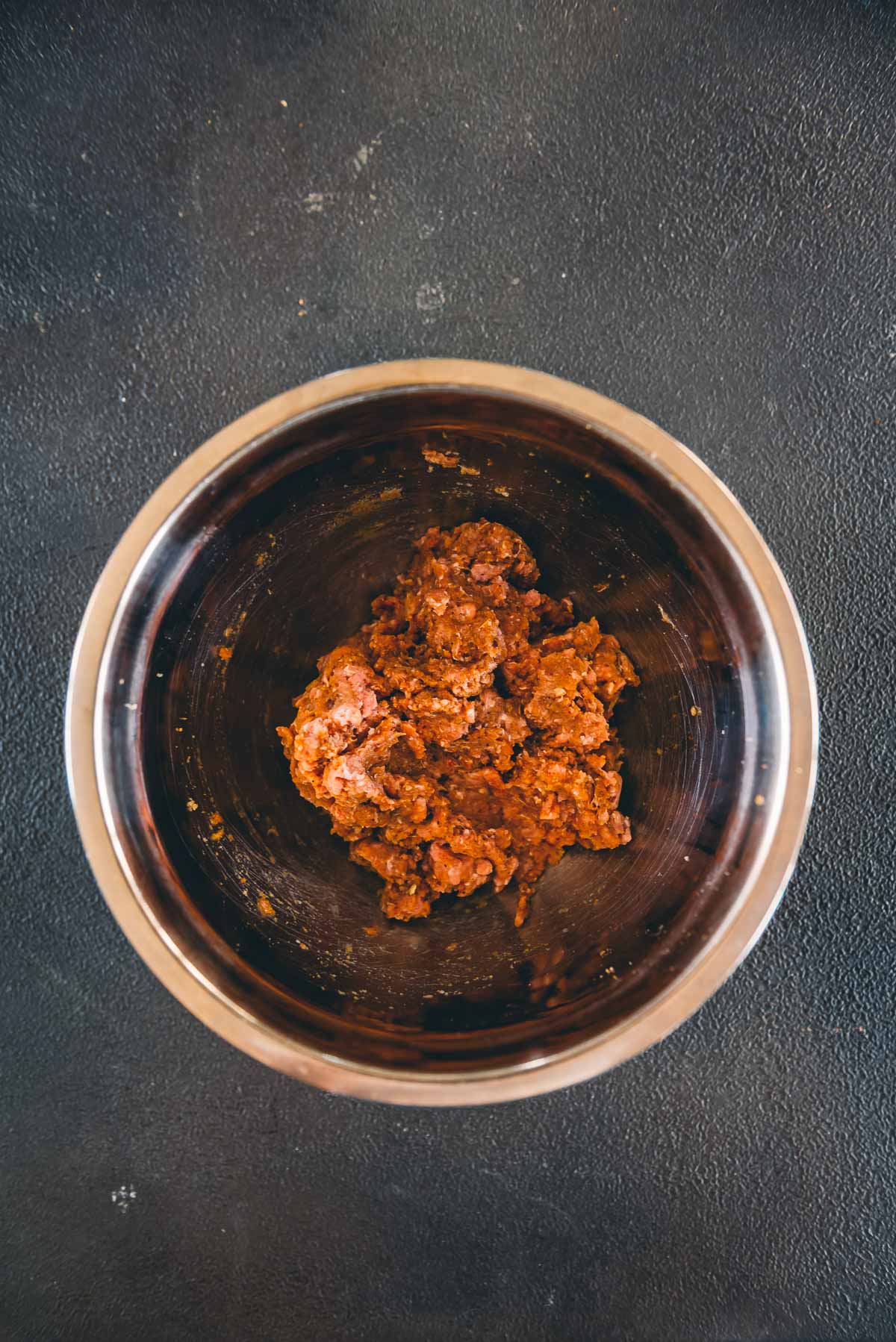 A stainless steel bowl containing a portion of seasoned raw ground turkey sits on a dark textured surface.