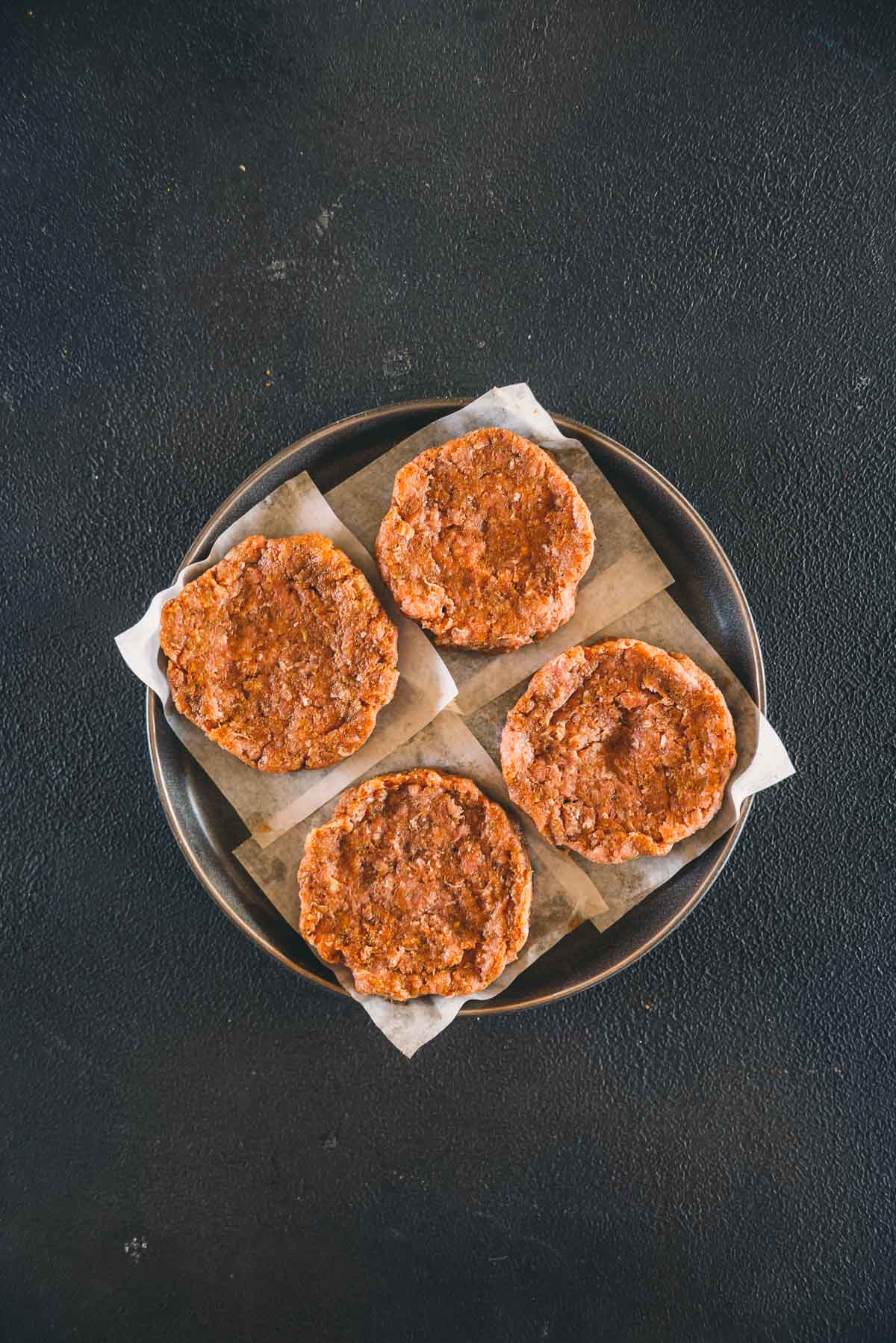 Four round, turkey burger patties are arranged on parchment paper squares on a round plate, set against a dark textured background.
