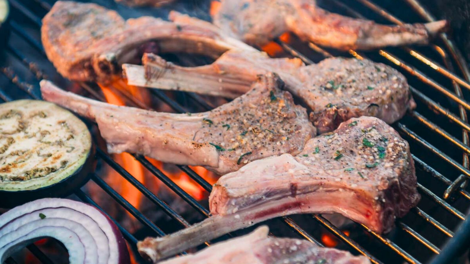 Lamb chops and sliced vegetables, including eggplant and red onion, are grilling over an open flame on a barbecue.