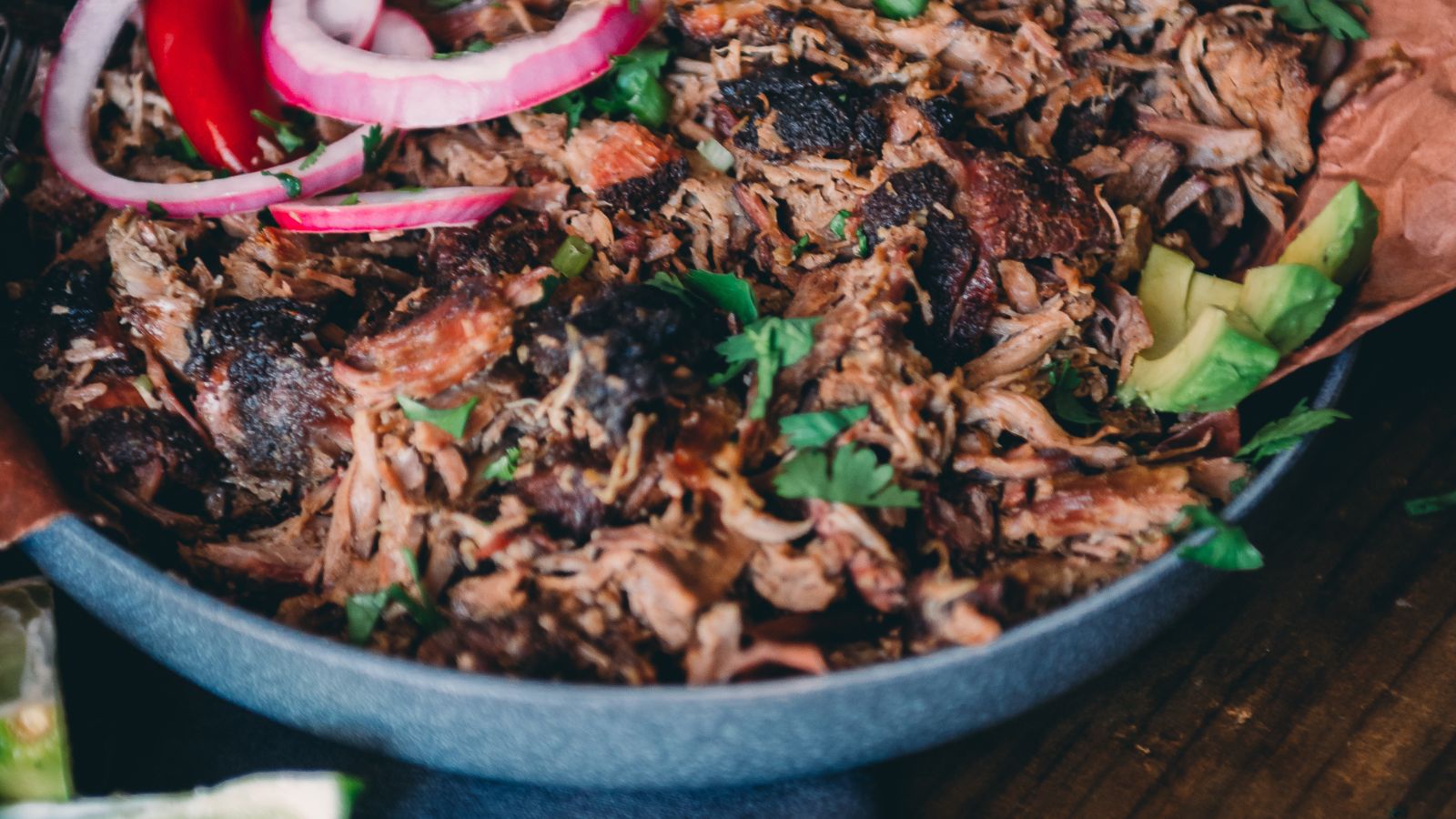 A close-up of a bowl filled with shredded barbecued meat, garnished with sliced red onions, avocado, and fresh herbs.