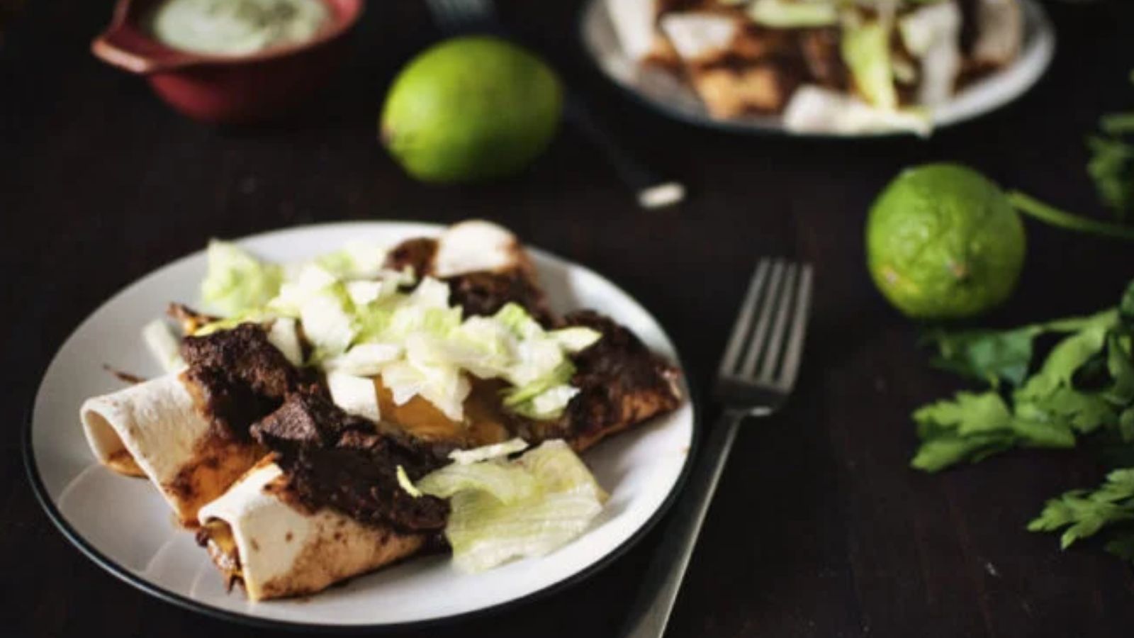 A plate with three enchiladas topped with chopped lettuce, a fork, two limes, and fresh cilantro on a dark surface. Another plate and a bowl of sauce are in the background.
