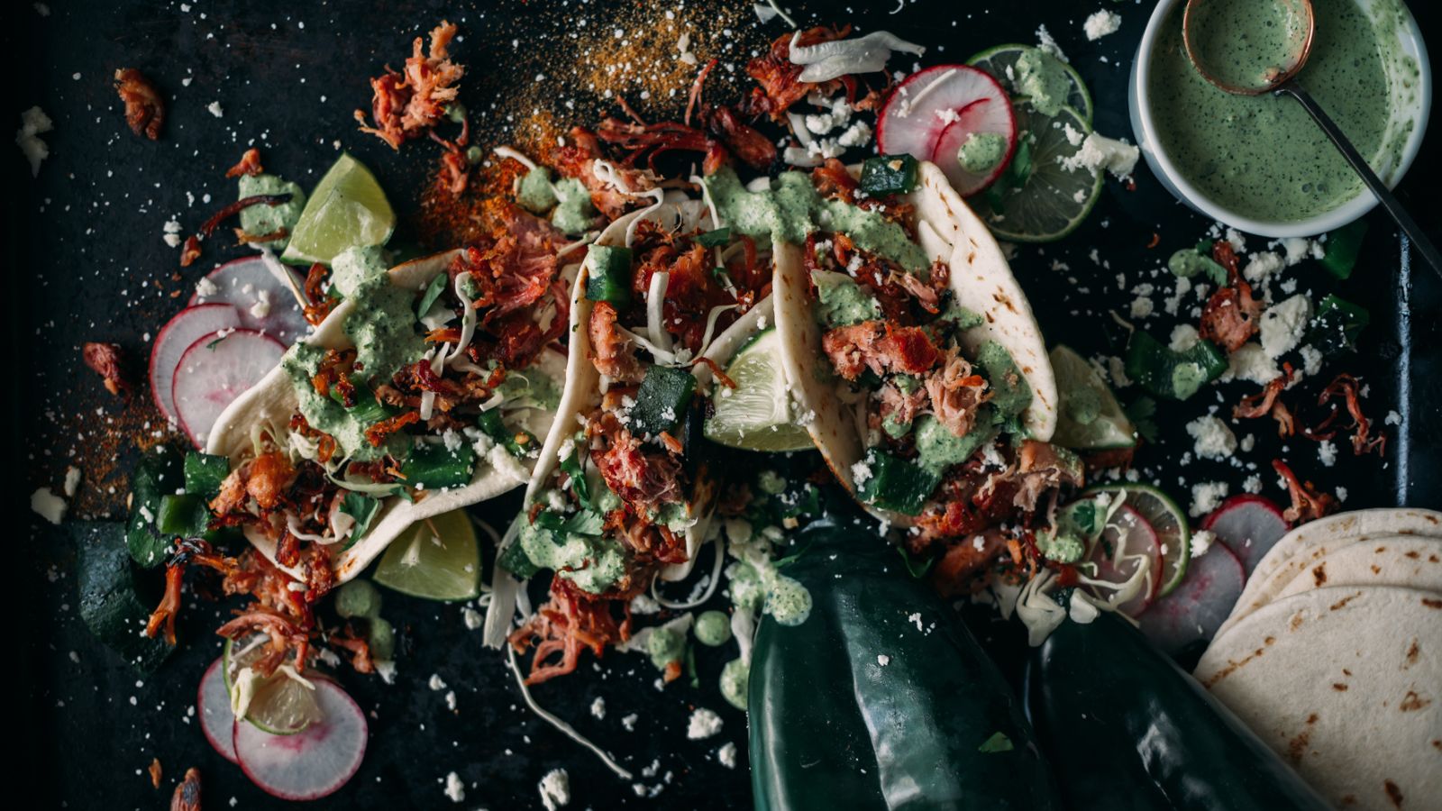 Three tacos filled with shredded meat, green sauce, and toppings are arranged on a dark surface with lime wedges, radish slices, cheese crumbles, tortillas, and poblano peppers scattered around.