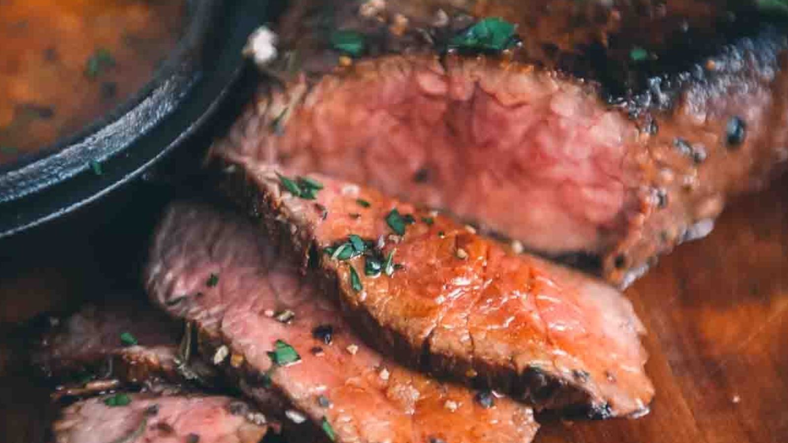 Sliced medium-rare steak garnished with herbs on a wooden surface, with a bowl of sauce partially visible.