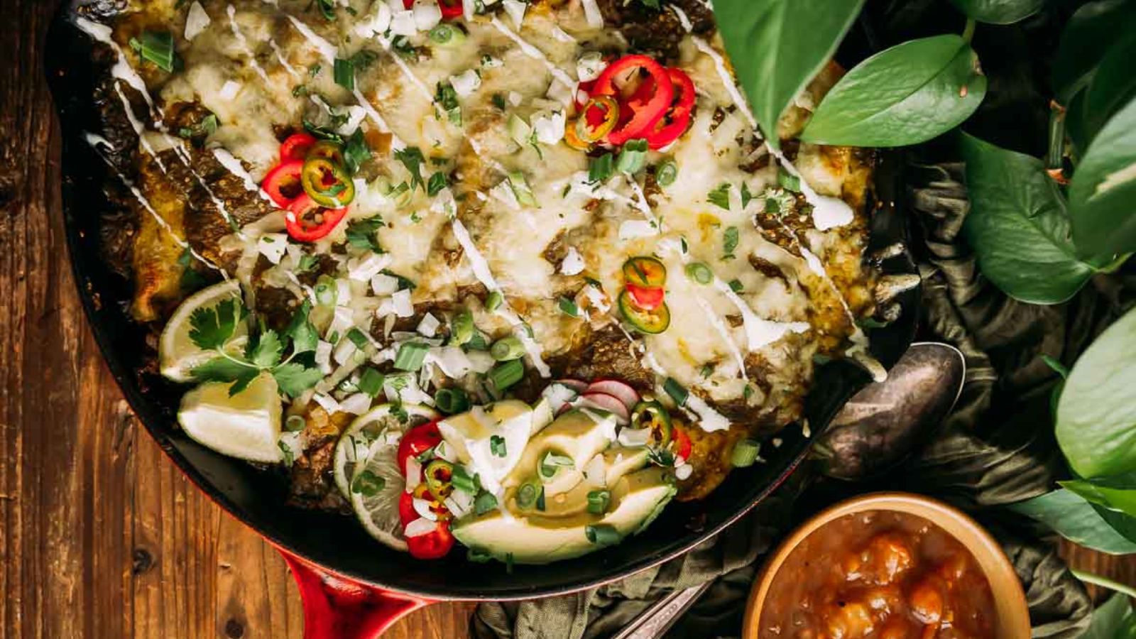 A skillet filled with baked enchiladas topped with melted cheese, sliced peppers, onions, cilantro, and lime wedges, with a bowl of salsa on the side.