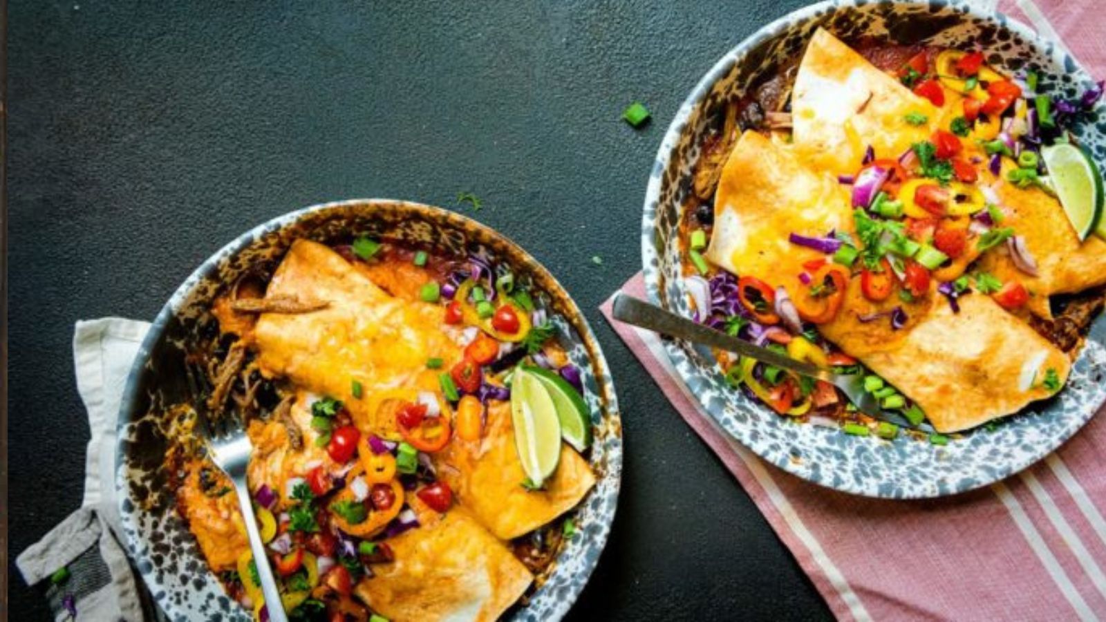 Two bowls of enchiladas topped with melted cheese, chopped vegetables, and lime wedges sit on a dark surface with a striped napkin beside them.