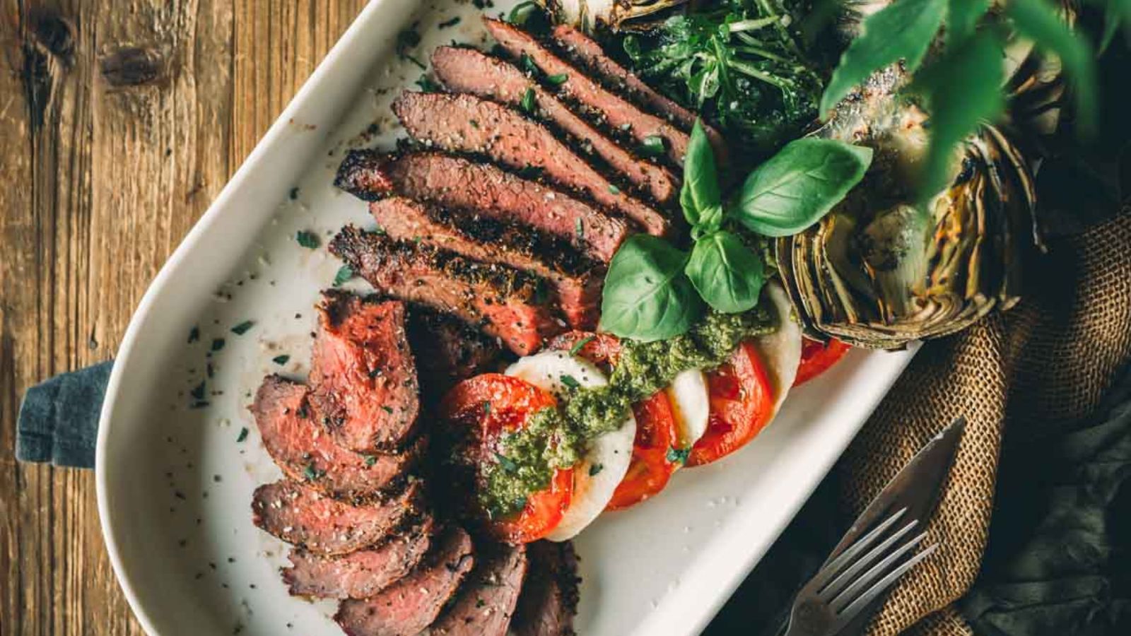 Sliced smoked tri-tip with tomato and mozzarella salad, topped with pesto and garnished with basil, served on a white platter with greens and artichoke.