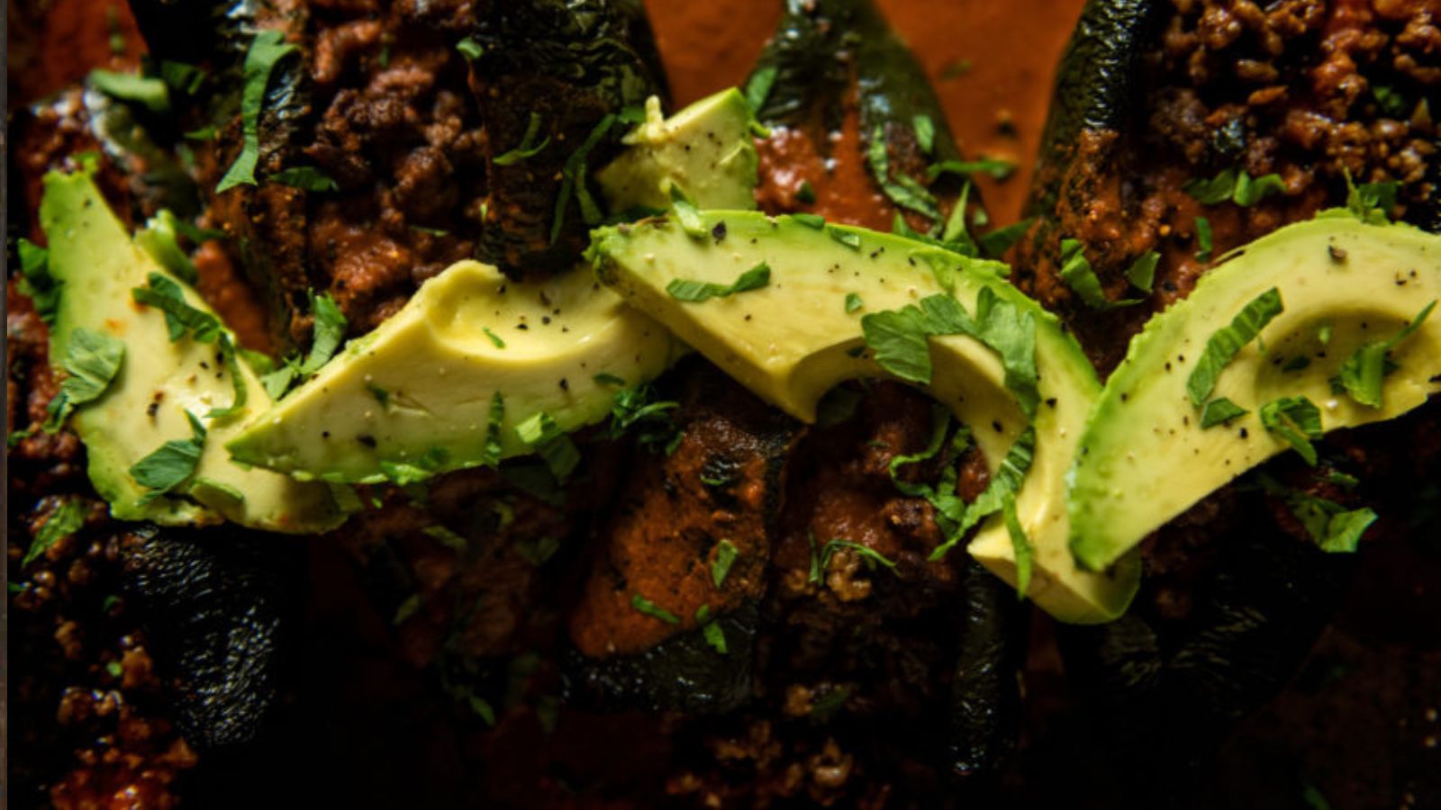 Close-up of stuffed poblano peppers topped with ground meat, avocado slices, and chopped cilantro.