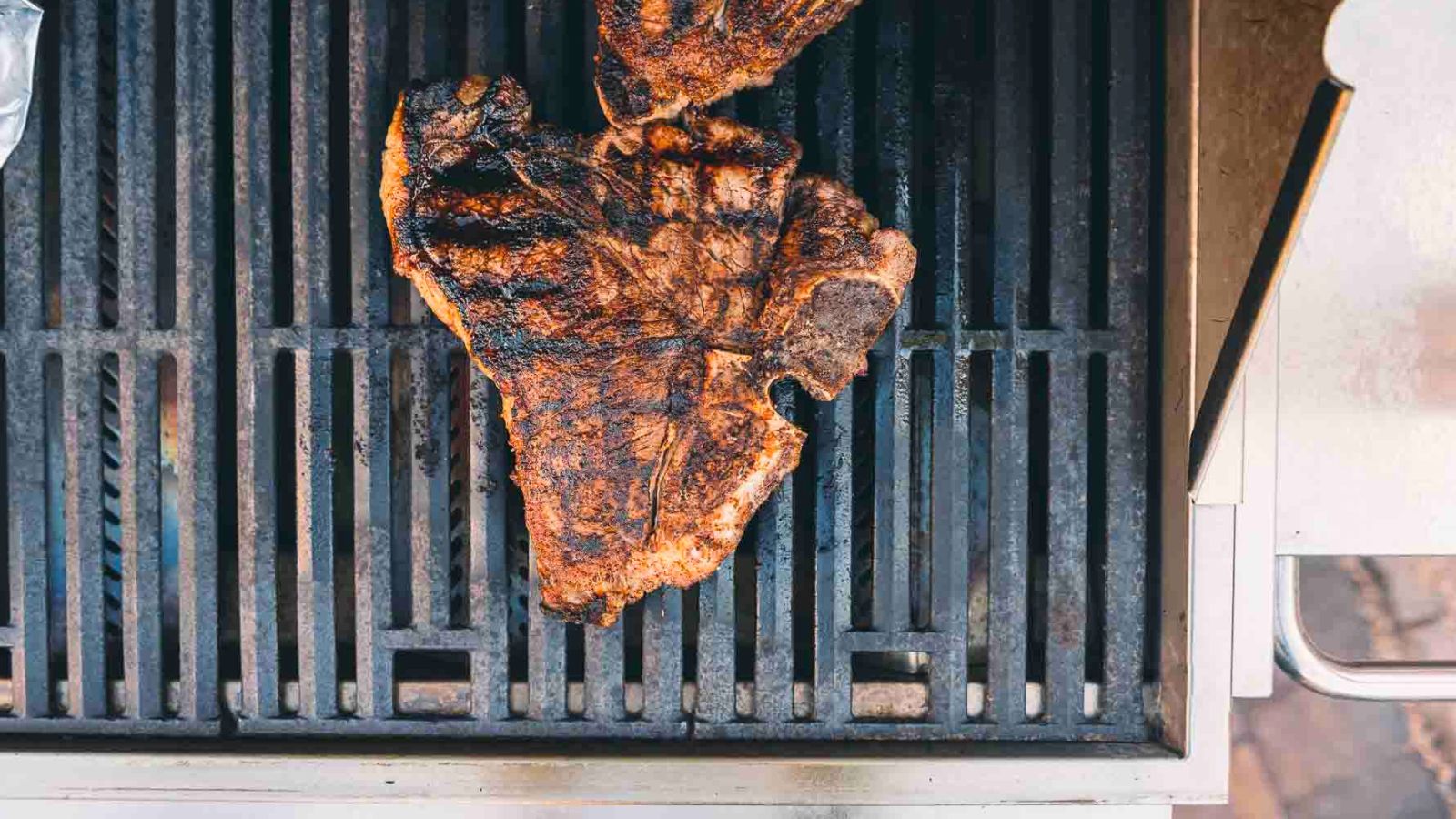 A large, grilled T-bone steak with char marks sits on a metal grill.