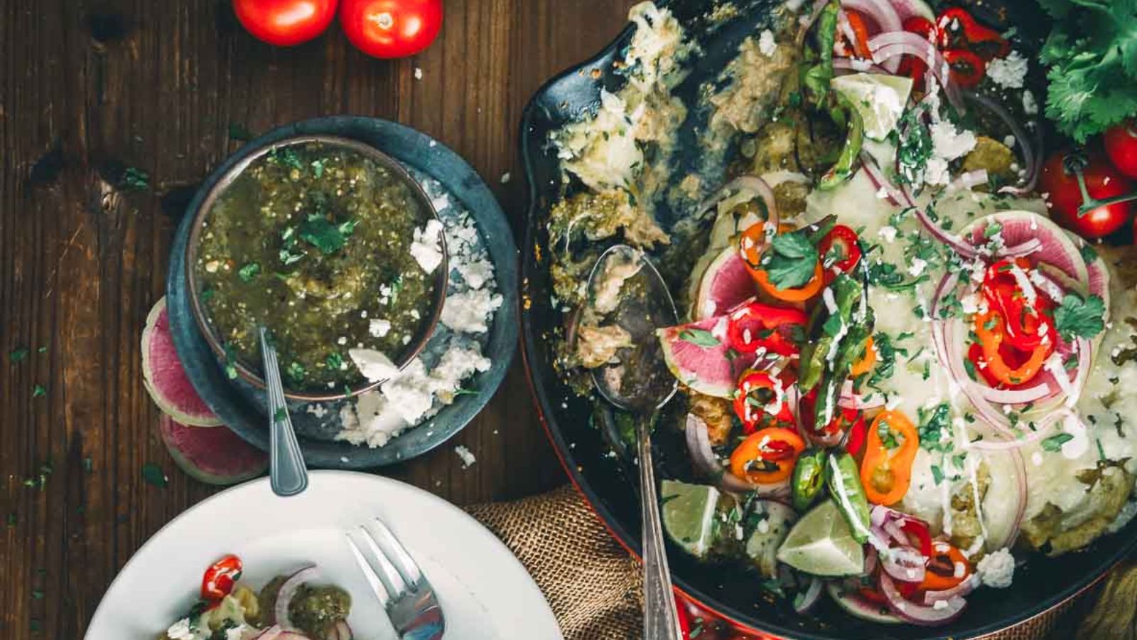 A skillet of enchiladas topped with onions, peppers, and cilantro beside a bowl of green salsa, tomatoes, and a plate with a serving of the dish on a wooden table.