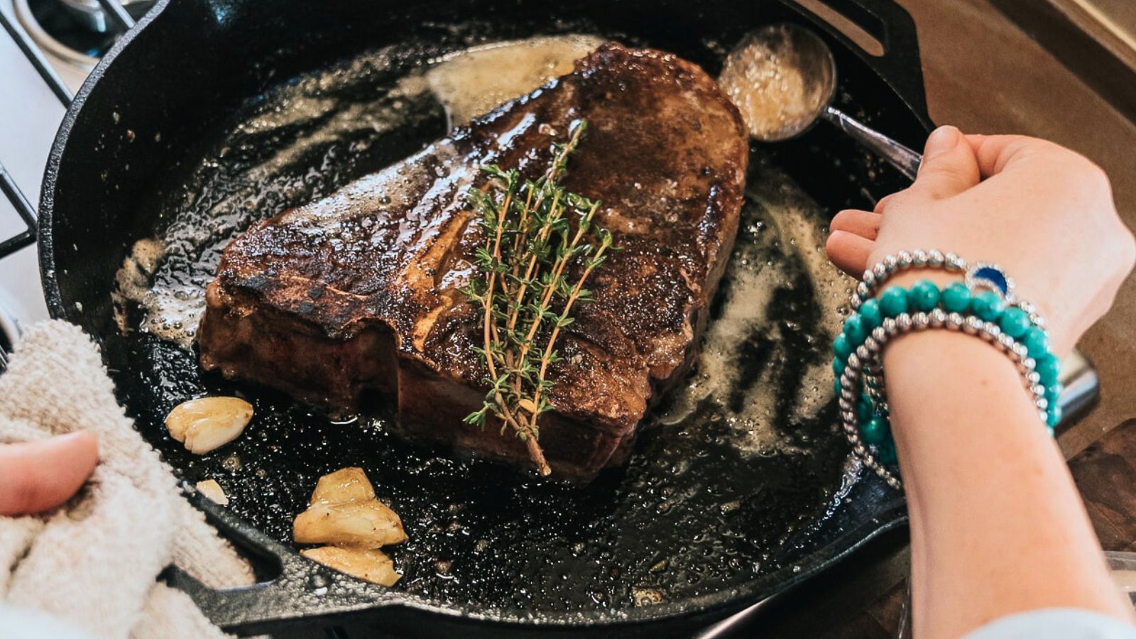 A person spoons melted butter over a seared steak with herbs and garlic in a cast iron skillet on a stovetop.