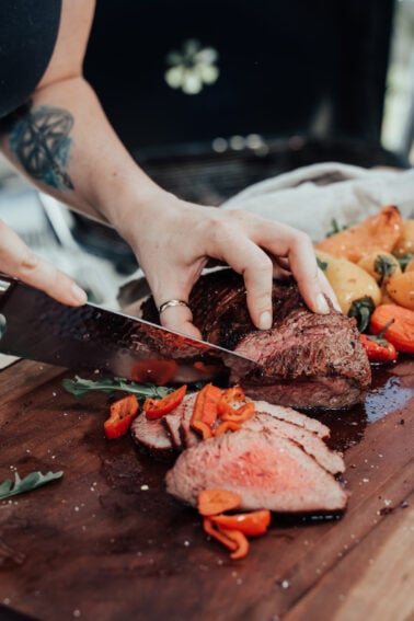 A person slices a cooked roast beef on a wooden cutting board, surrounded by grilled vegetables such as peppers and tomatoes.