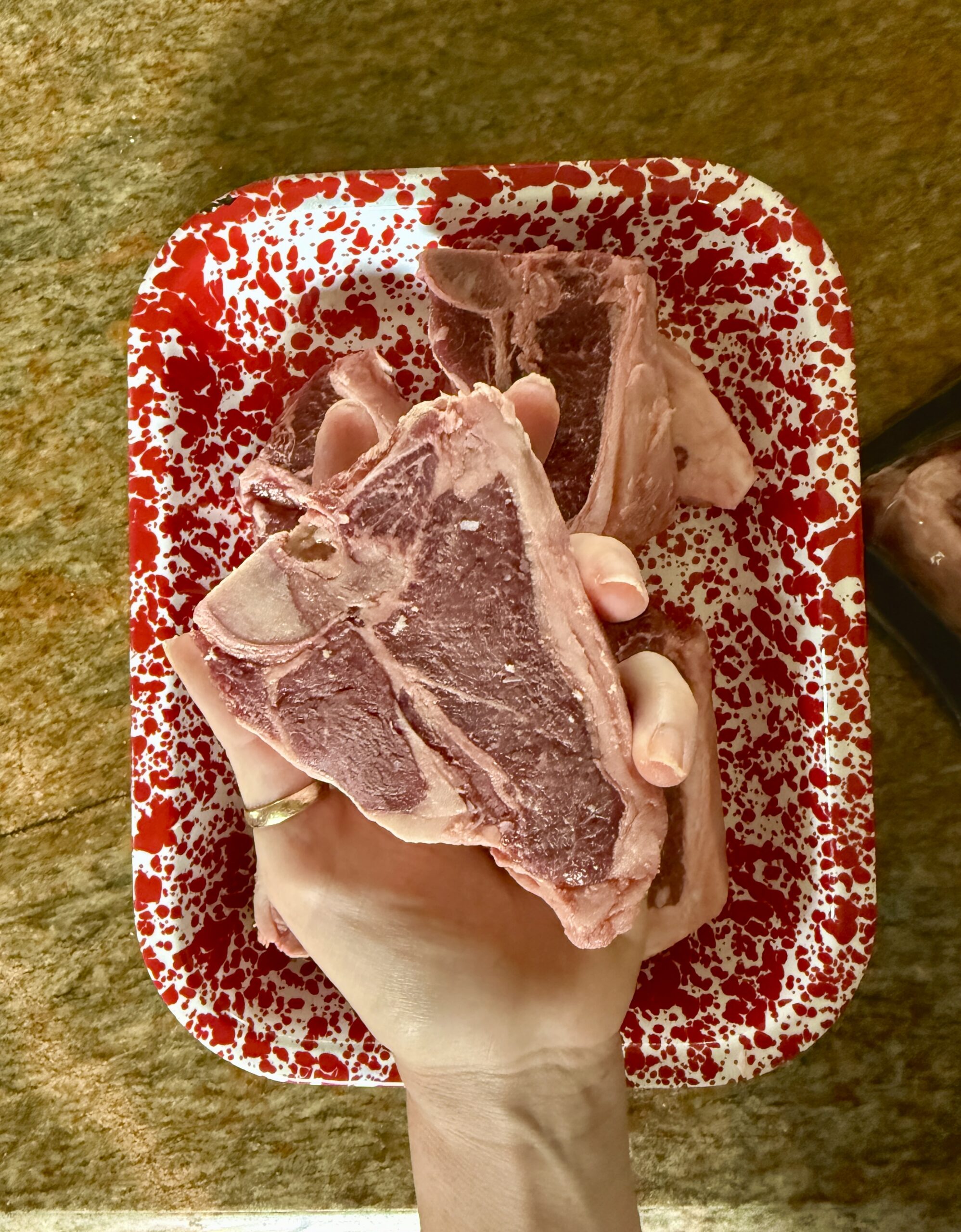 A hand holding a raw T-bone steak over a red and white speckled tray with more T-bone steaks and grilled lamb loin chops, all set on a brown countertop.