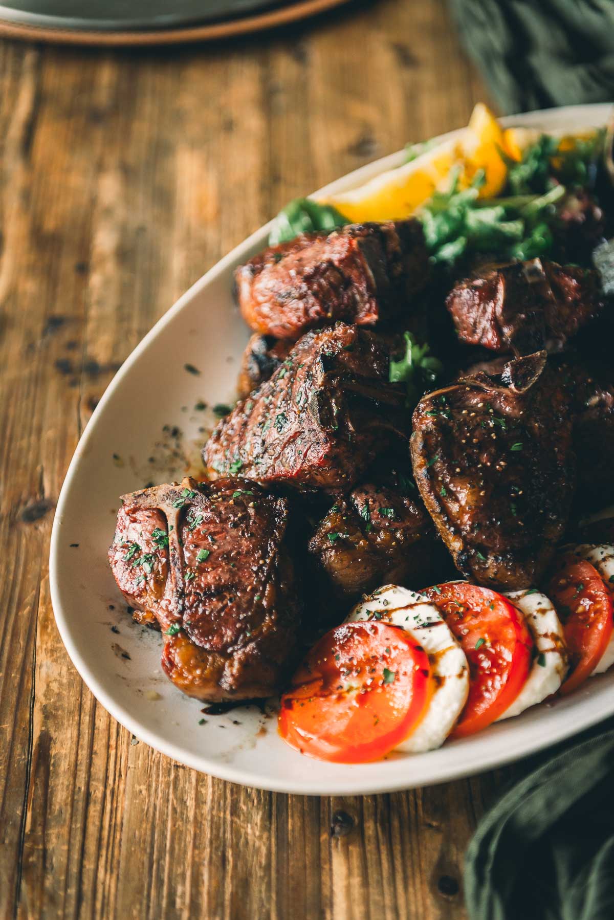 A plate of grilled lamb loin chops garnished with herbs, served with sliced tomatoes, mozzarella, and greens on a rustic wooden table.