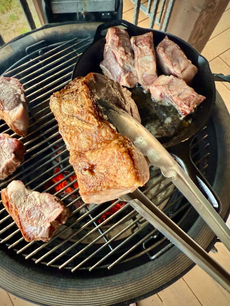 A pair of tongs holds a browned piece of beef over a grill with several other cuts, including grilled lamb loin chops, sizzling nearby—some are searing in a cast iron pan above red-hot coals.