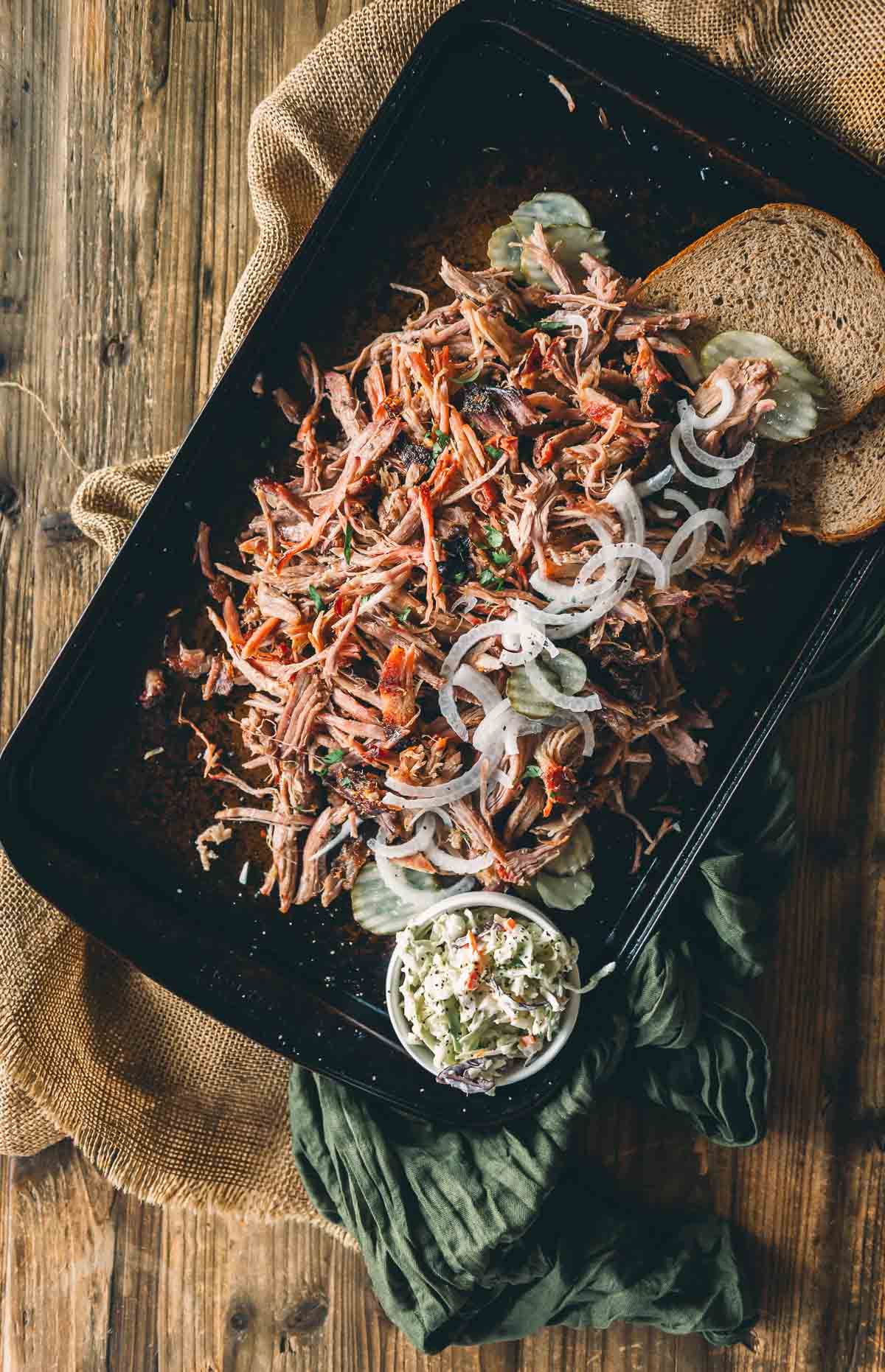 A tray of shredded smoked picnic shoulder topped with sliced onions, served with rye bread, pickle slices, and a side of coleslaw on a rustic wooden table.
