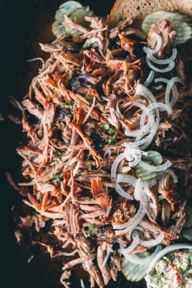 Pulled pork served on a slice of bread, topped with sliced pickles and raw onions, with a side of coleslaw partially visible in the corner.