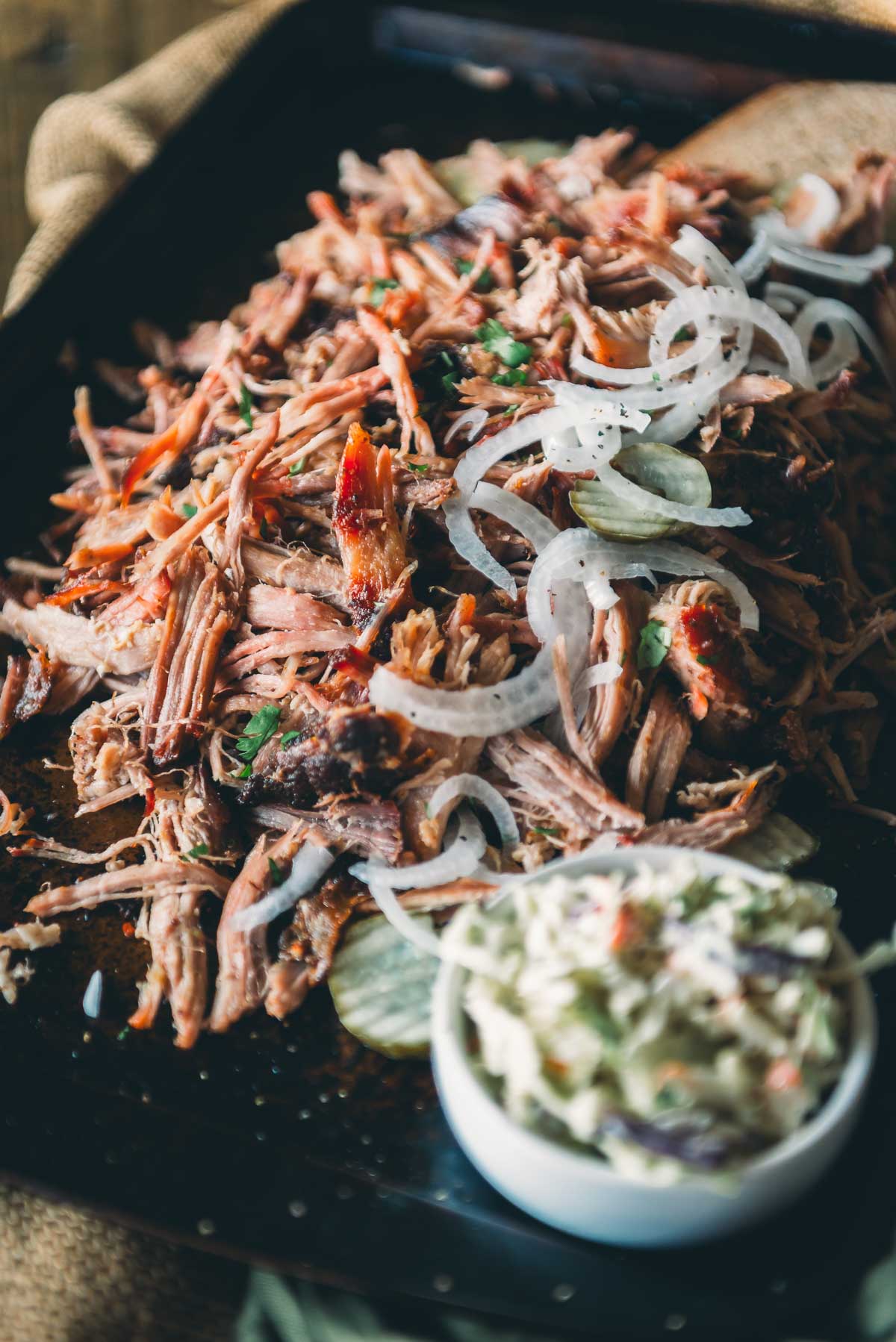 A tray of pulled pork garnished with sliced onions and herbs, with a small bowl of coleslaw on the side.
