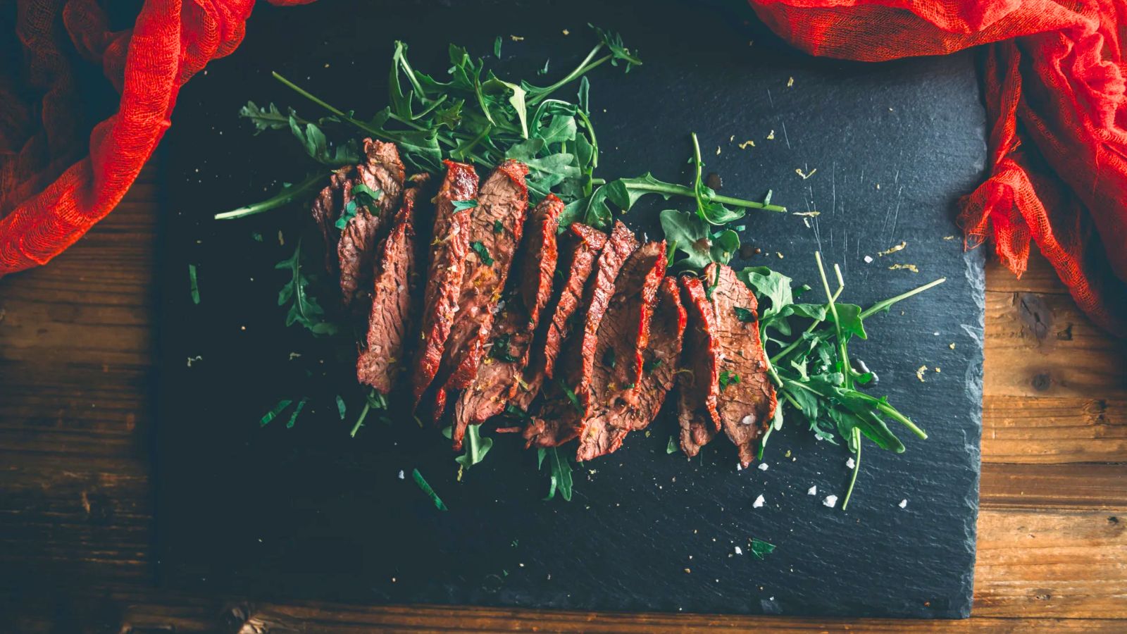 Sliced grilled steak arranged on a bed of arugula leaves atop a black slate serving board, with a red cloth partially visible in the corners.