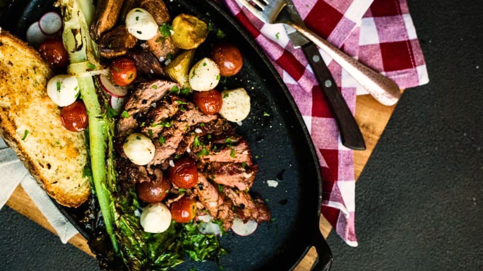 A plate of grilled steak slices, cherry tomatoes, potatoes, mozzarella balls, lettuce, and toast on a wooden board with a red checkered napkin and cutlery.