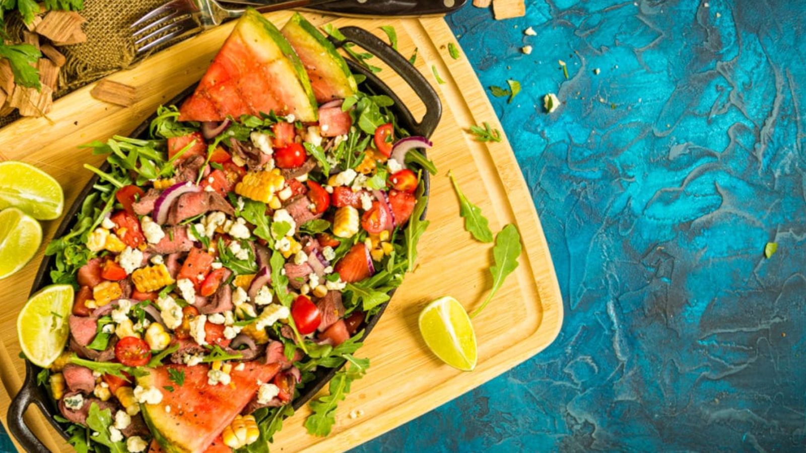 A platter of watermelon salad with arugula, cherry tomatoes, red onion, feta cheese, and lime wedges on a wooden board against a blue textured background.