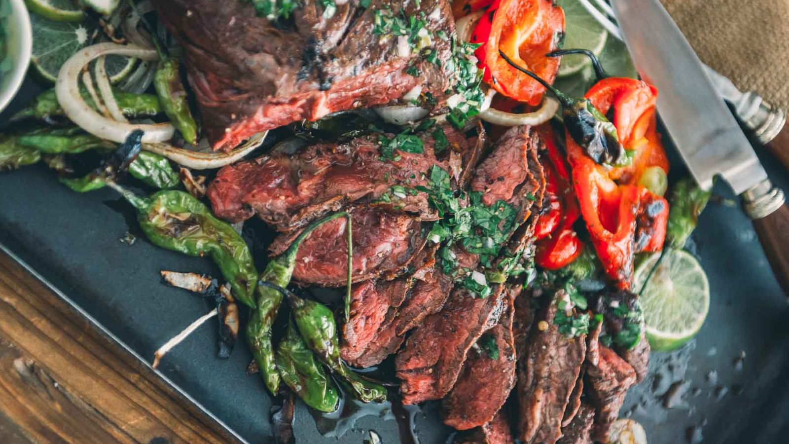 Sliced grilled steak served with roasted peppers, onions, lime slices, and herbs on a black platter, with a knife and fork beside the food.