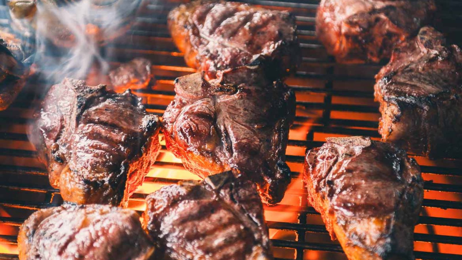 Several pieces of steak are being grilled over an open flame, with visible grill marks and smoke rising.