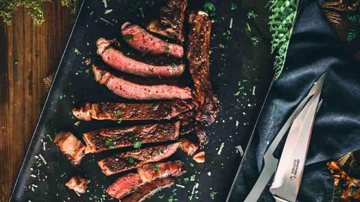 Sliced grilled steak arranged on a dark cutting board, garnished with chopped herbs, with a knife and fork placed nearby.