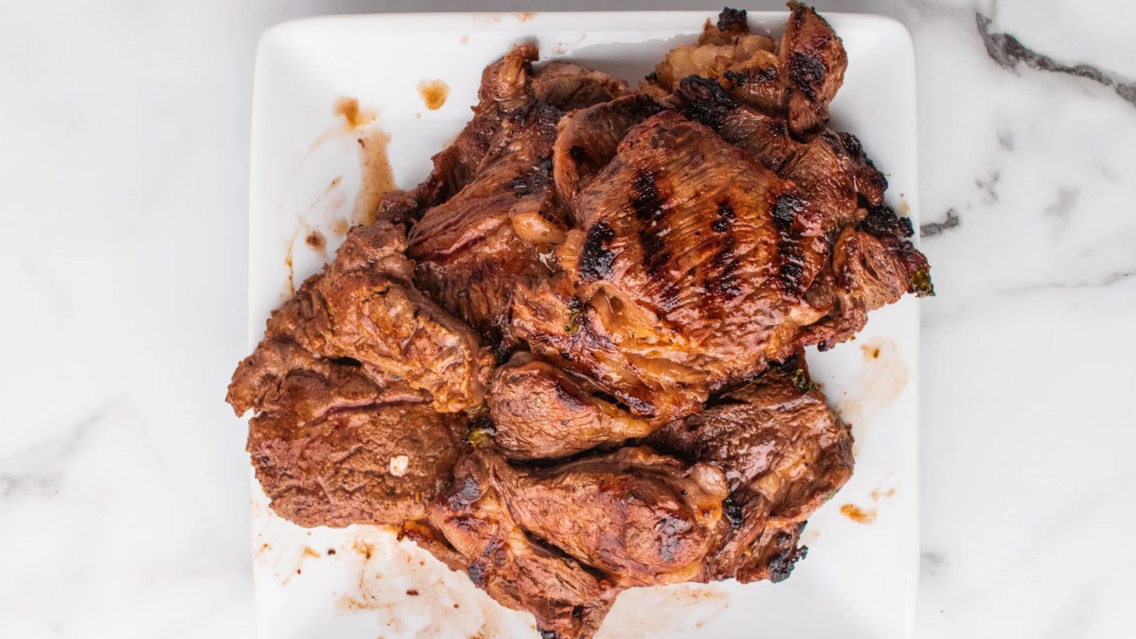 Grilled, marinated pieces of steak are stacked on a white square plate placed on a white marble surface.