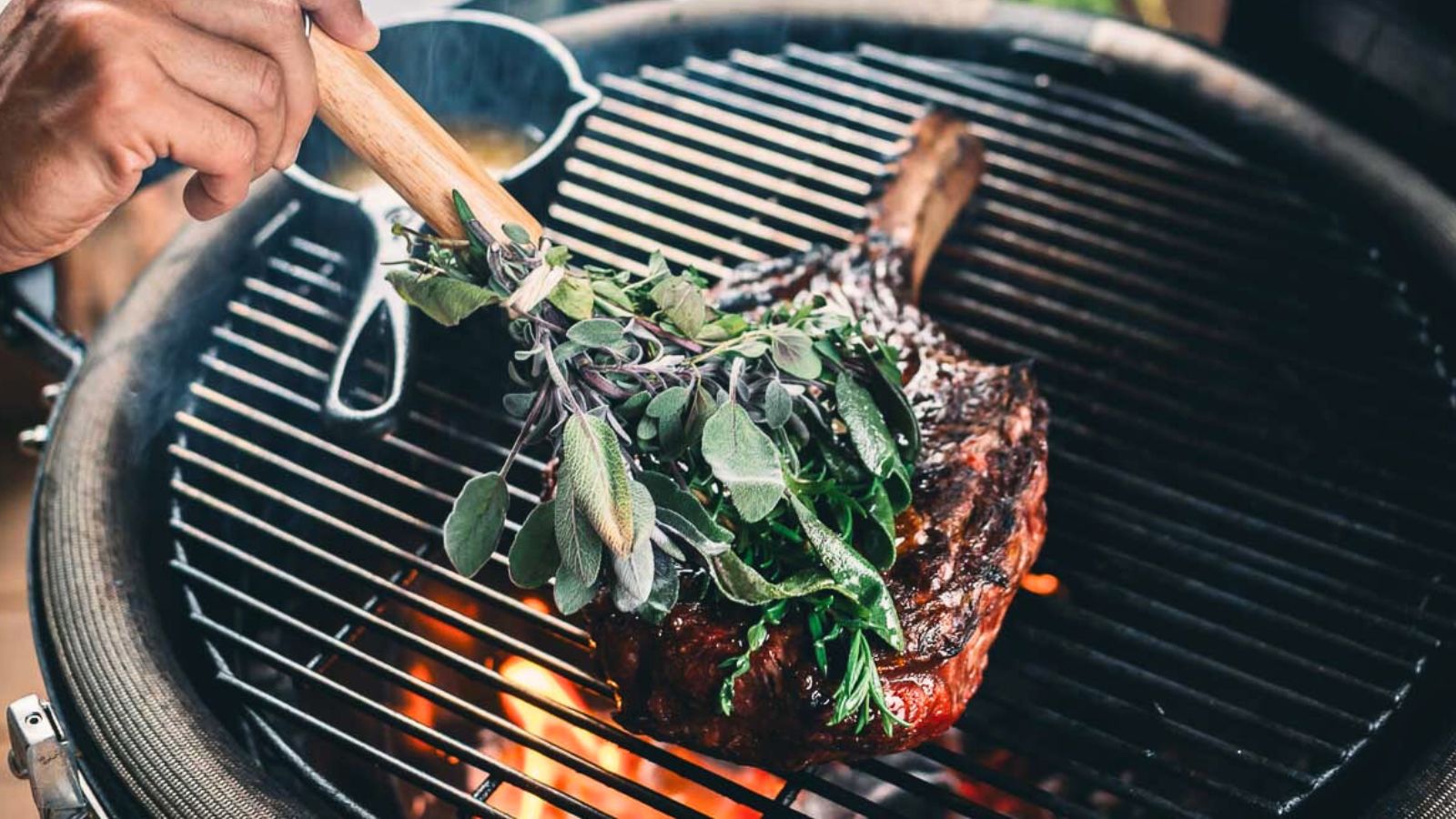 A hand uses a bunch of fresh herbs tied to a wooden handle to baste a large steak cooking on a grill over open flames.