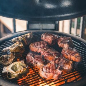 Lamb loin chops and artichoke halves are grilling over an open flame on a round, red barbecue grill with the lid open.
