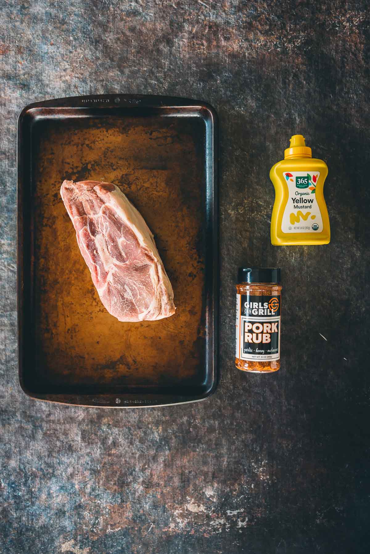 Raw pork picnic shoulder on a sheet tray next to yellow mustard and pork rub.