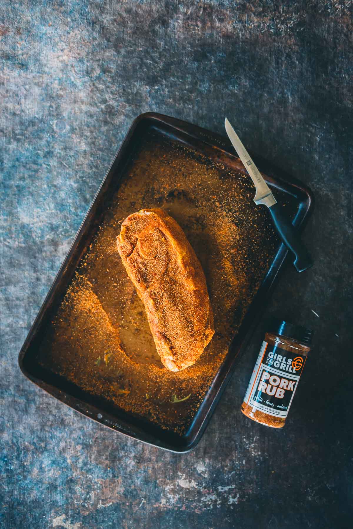 A seasoned pork roast sits on a baking tray with a fillet knife and a container of pork rub seasoning beside it.