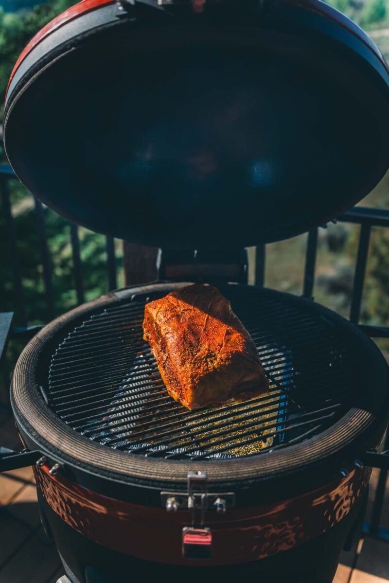 A seasoned pork roast sits on the grill grate inside an open kamado-style charcoal grill outdoors.