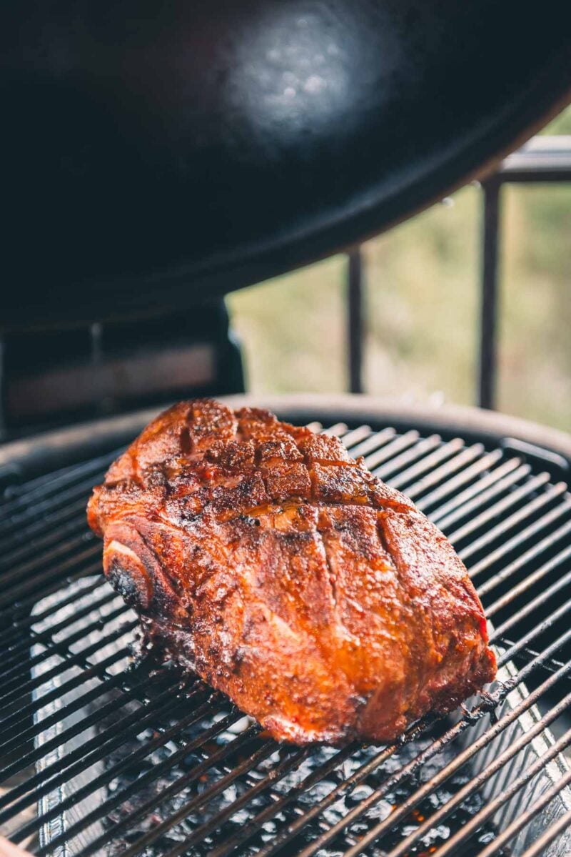 A seasoned pork picnic roast is being smoked on a charcoal grill with the grill lid open.