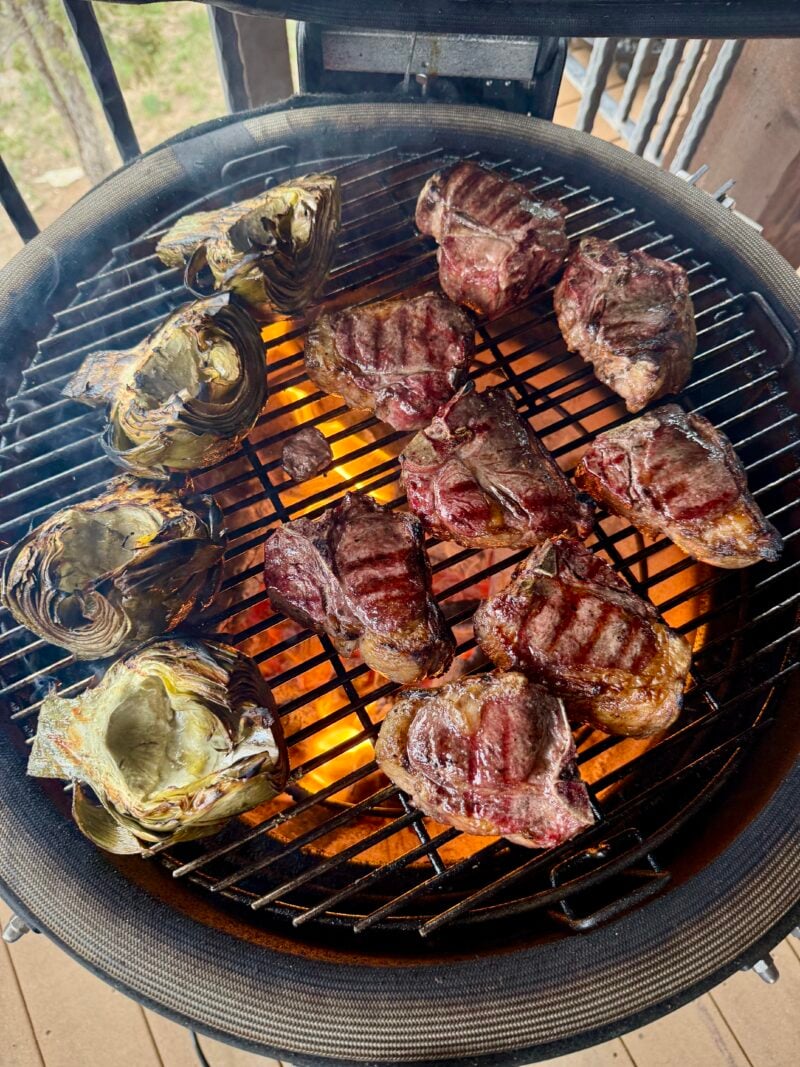 Several pieces of grilled steak, grilled lamb loin chops, and halved artichokes cook over an open flame on a round outdoor grill.