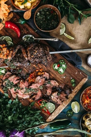 Overhead view of a rustic table filled with grilled meat on a wooden board, guacamole, chips, salsa, herbs, vegetables, and various side dishes.