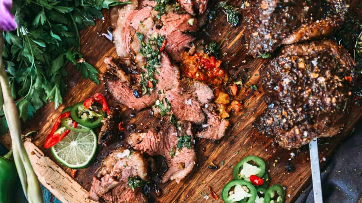 Sliced grilled steak on a wooden board with herbs, lime, sliced jalapeรฑos, and chopped vegetables.