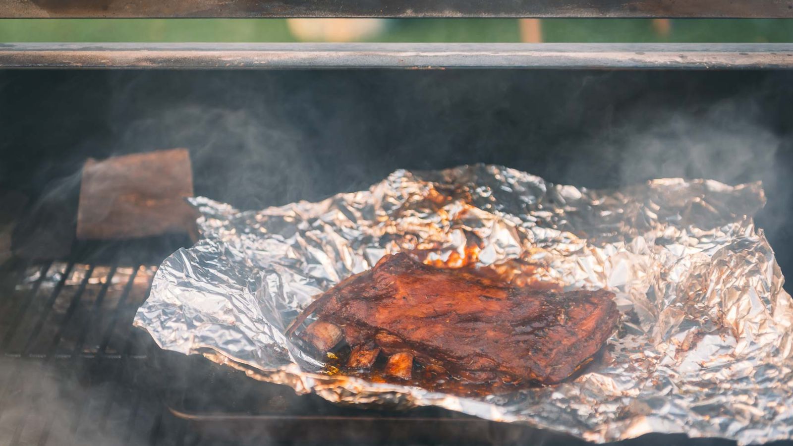 Barbecued meat resting on aluminum foil inside a smoker, with smoke swirling around and a grill grate visible underneathโperfect inspiration for your next Fathers Day recipes.